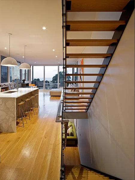 Interior view of a modern, open-plan apartment with a staircase, kitchen island, and large windows.