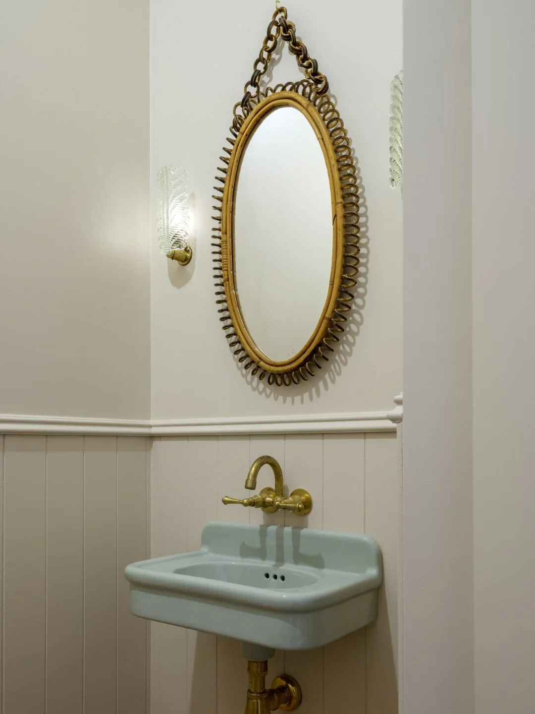 An oval mirror with a rattan frame and chain hangs above a vintage green wall-mounted sink with brass fixtures, against a white wall with a wall-mounted light.