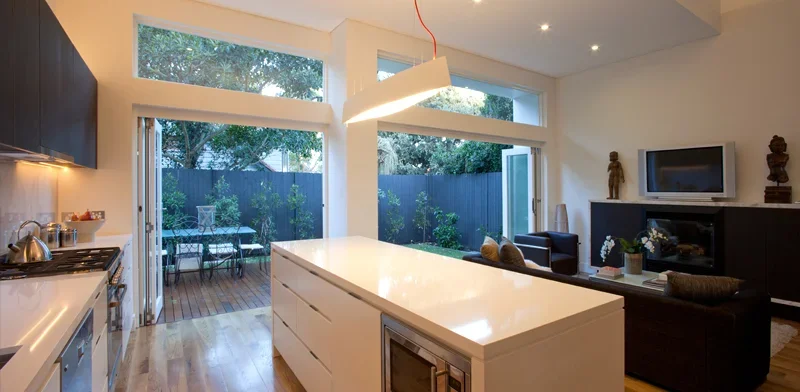 Modern open-concept kitchen and living room with large windows overlooking a backyard patio with outdoor furniture and trees.