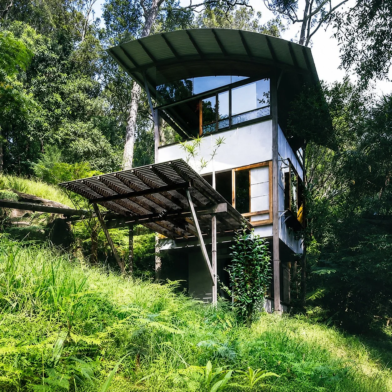 Designed by Peter Stutchbury. Unique multi-level house with a green curved roof, elevated on stilts, surrounded by lush greenery in a forest setting.