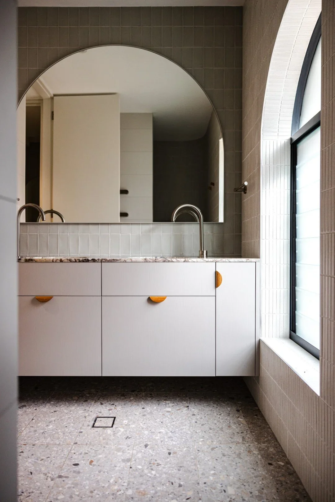 A minimalist bathroom with a large mirrored arch, white cabinet with wooden handles, and a window with frosted glass.