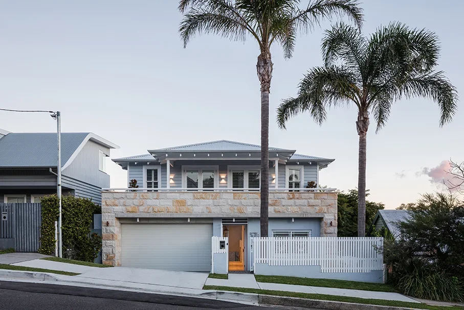 A modern two-story house with a light gray exterior, stone accents, and a white picket fence, surrounded by palm trees