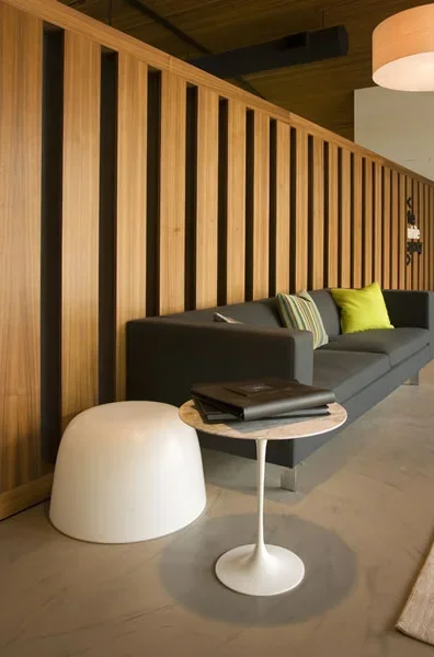Modern living room with a long dark gray sofa, green and gray pillows, a white round side table with a book, and a wooden-paneled wall.