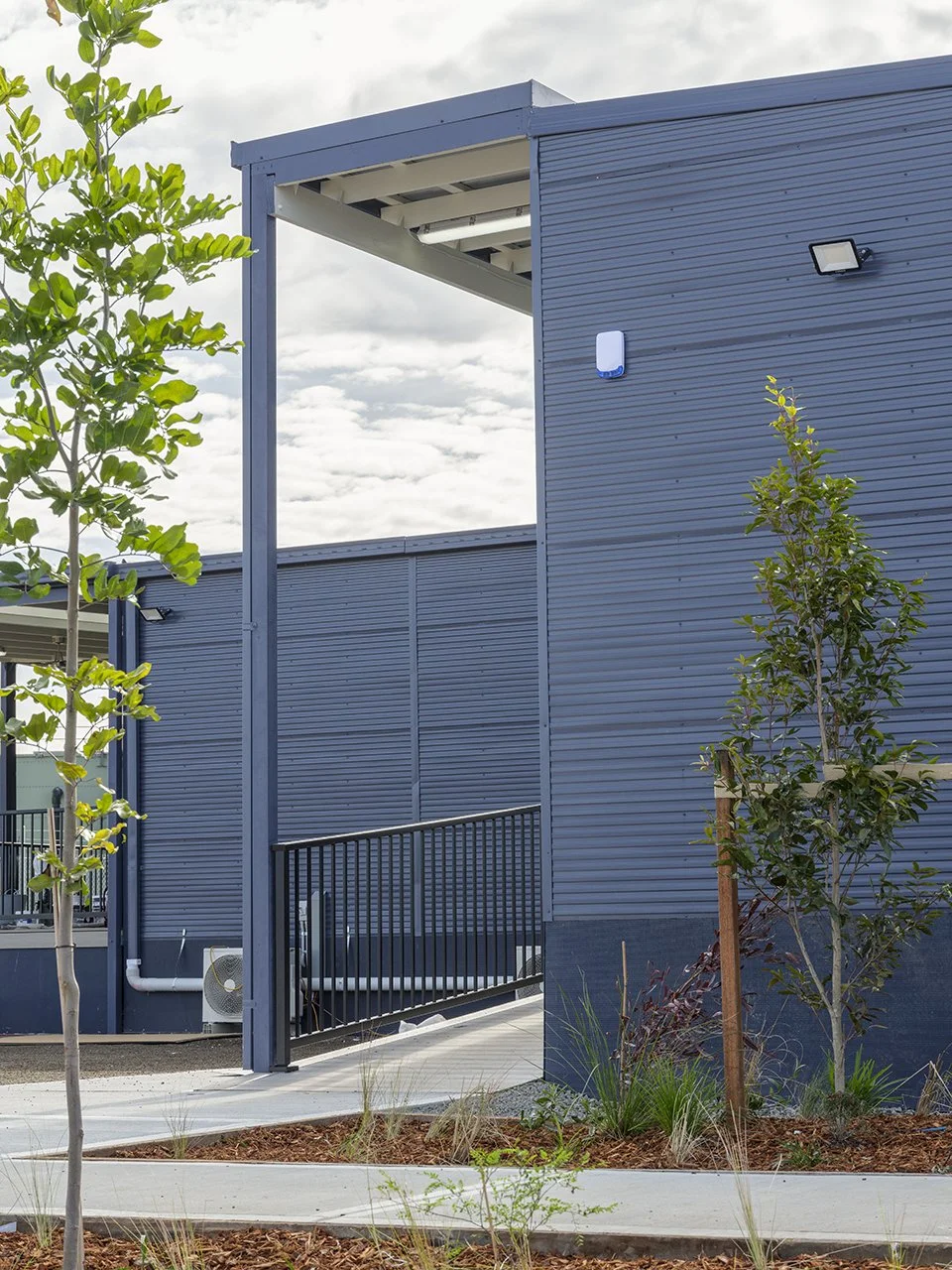 Modern blue building with metal siding, a small ramp, and landscaped plants nearby.