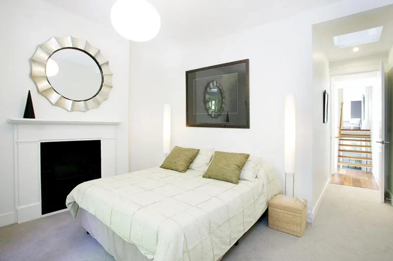 A modern bedroom with white walls, a bed with white bedding and two green pillows, a wicker storage box, wall-mounted lights, a fireplace, a decorative mirror above the fireplace, and a flat-screen TV on the wall. There is a doorway leading to a hall