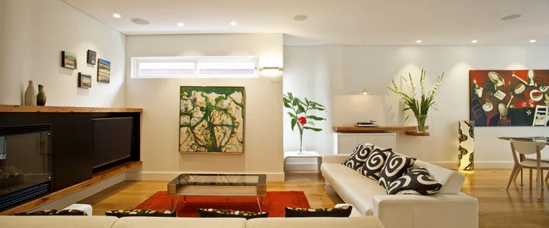 Modern living room with a white sofa, black and white patterned pillows, a glass coffee table, colorful artwork on the wall, a large plant, and a dining area in the background.