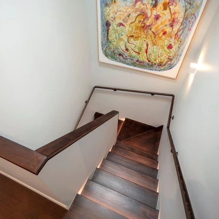 View down a staircase with wooden steps and dark wooden handrails on white walls. A colorful abstract painting hangs on the wall at the top of the stairs, and a wall-mounted light fixture illuminates the area.