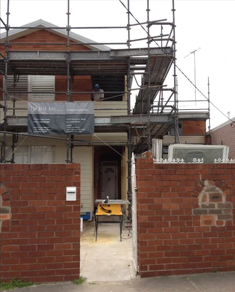 A house under construction with scaffolding around it, a yellow saw on a table in front, and a brick wall entrance.