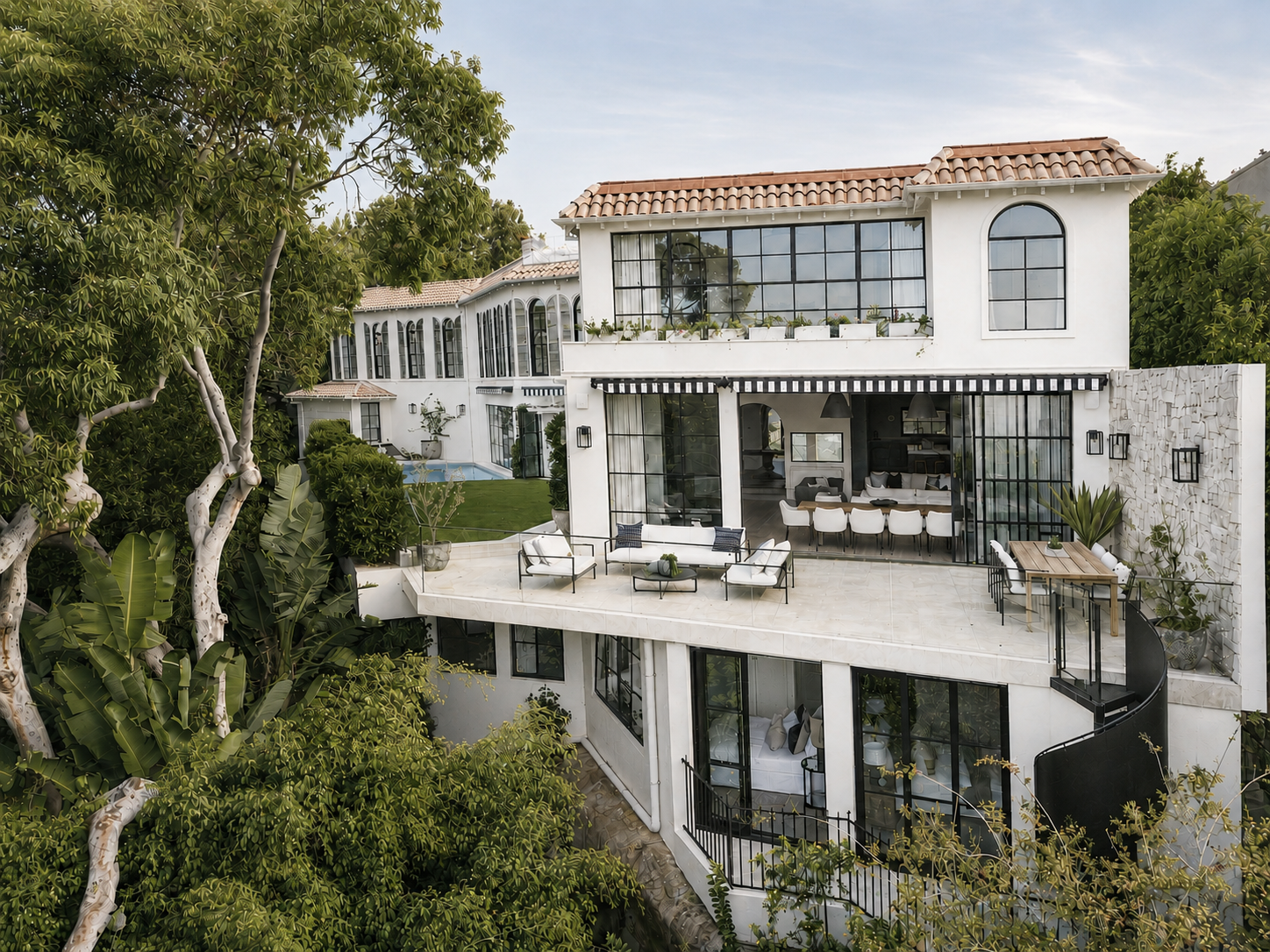 Modern multi-story white house with large glass windows and outdoor terrace, surrounded by trees and greenery.
