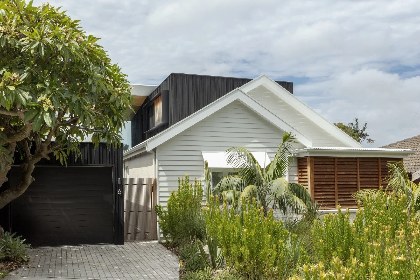 Modern house with white and black exterior, wooden accents, surrounded by green plants and trees, on a cloudy day.