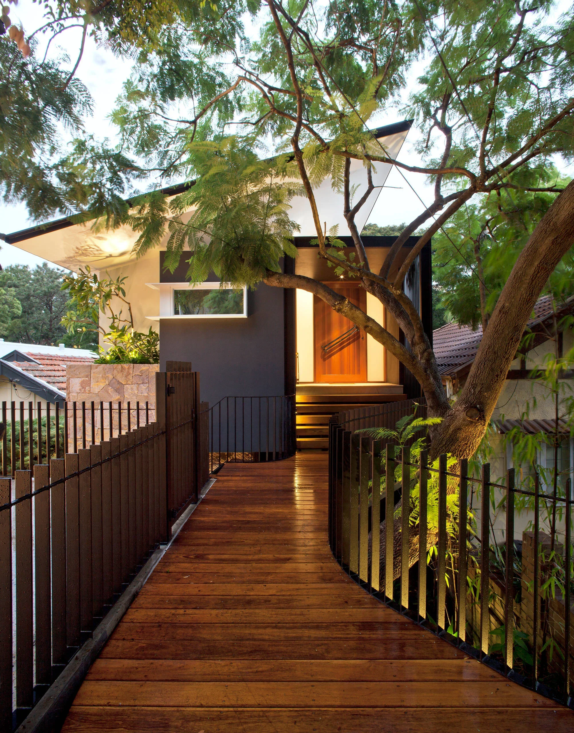 A modern house with a wooden walkway leading to the front door, surrounded by trees and greenery.