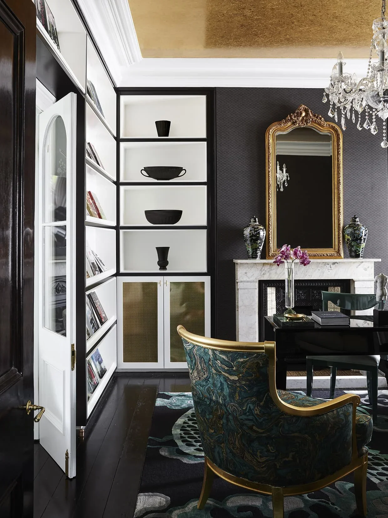 Elegant living room with a black and white built-in bookshelf, a gold-framed mirror above a white marble fireplace, and a chandelier. Furniture includes a floral-patterned armchair with gold trim and a dark dining table.
