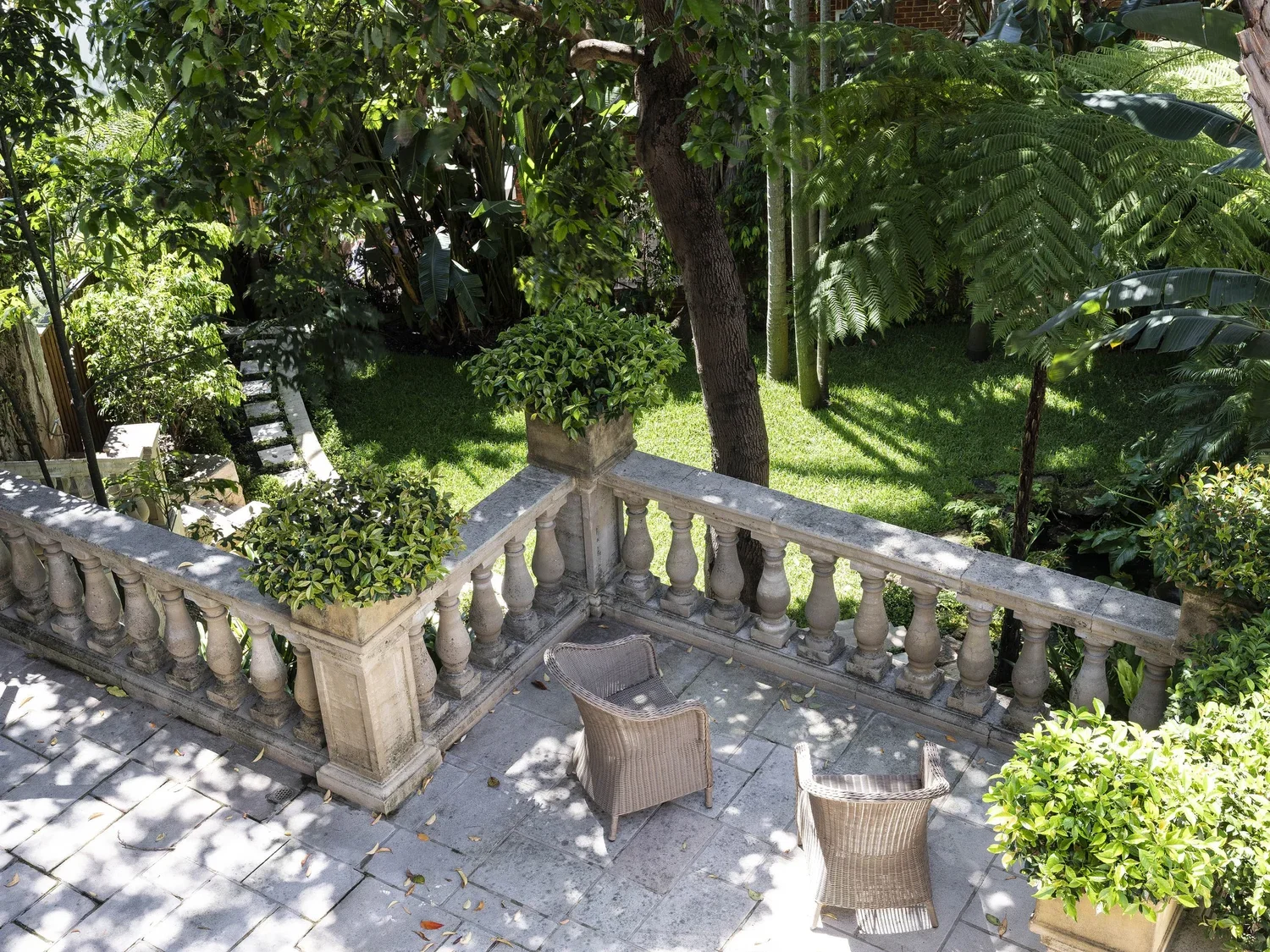 A garden patio with two wicker chairs and potted plants, surrounded by lush trees and greenery.
