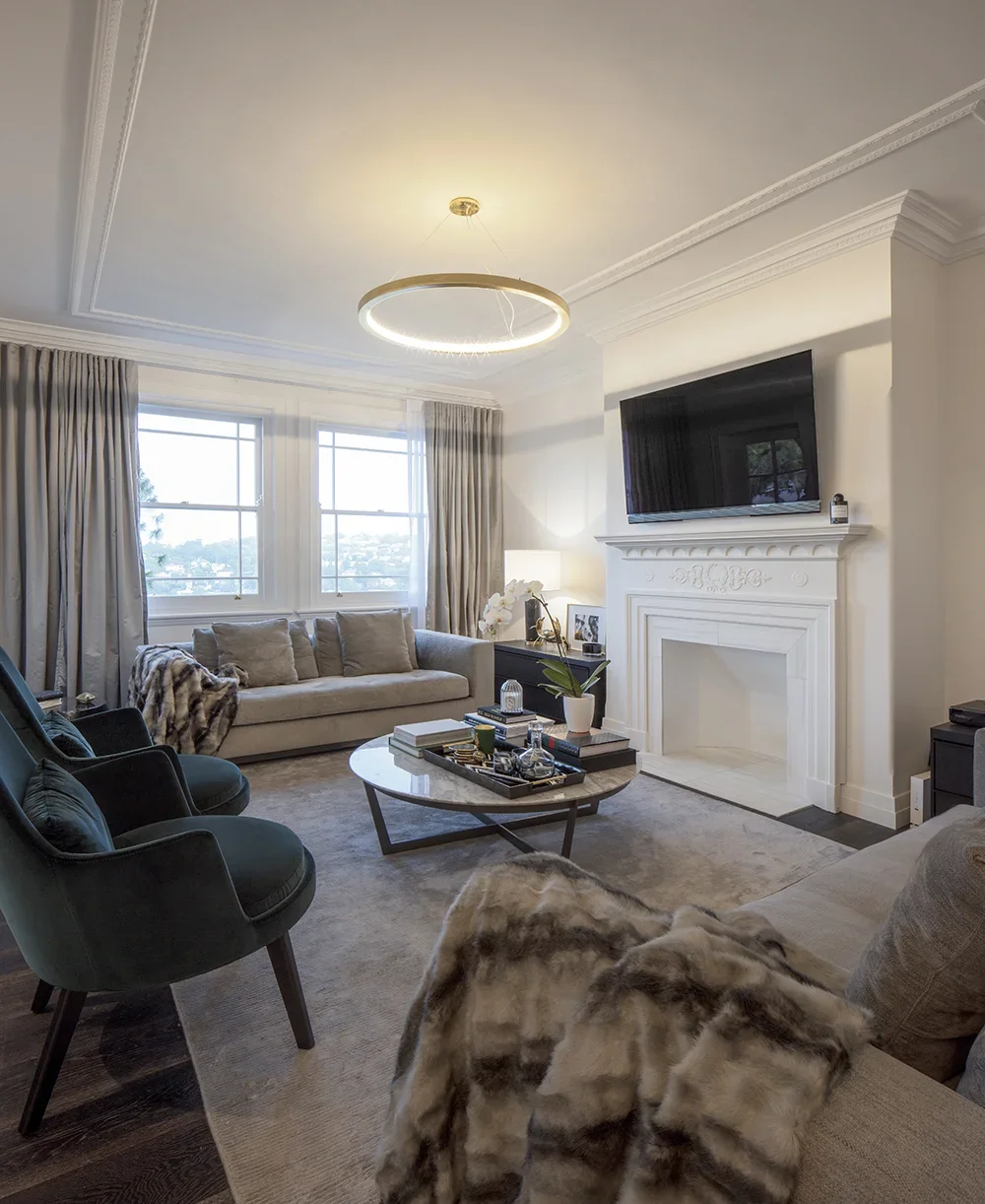 A cozy living room with large windows, a beige sofa, black armchairs, a marble coffee table, and a white fireplace with a mounted flat-screen TV above. Decorative pillows, a fur throw, and a side table with a lamp and orchid add to the inviting ambia