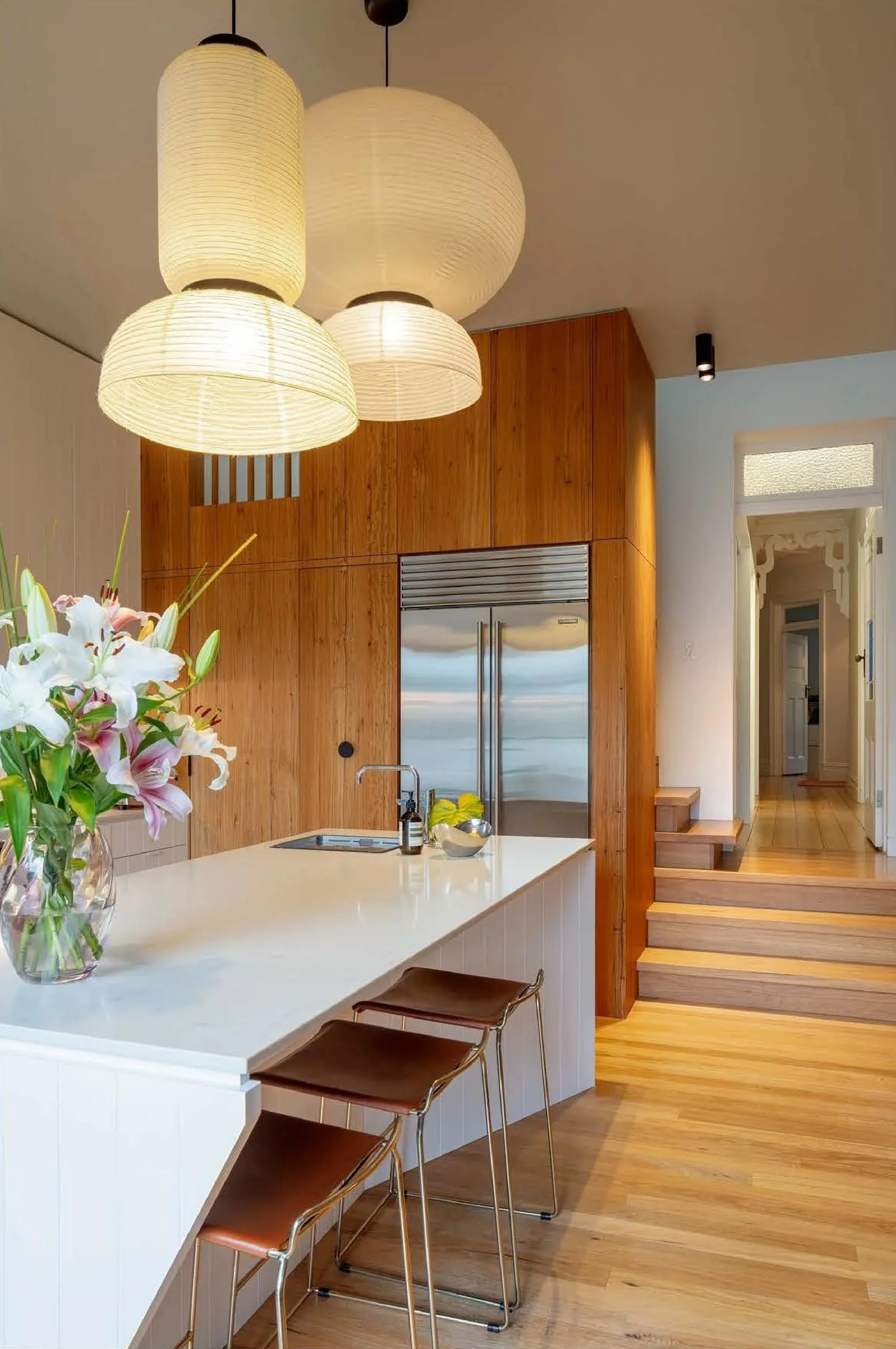 Modern kitchen with white island, wooden accents, stainless steel refrigerator, and hanging paper lantern light fixtures.