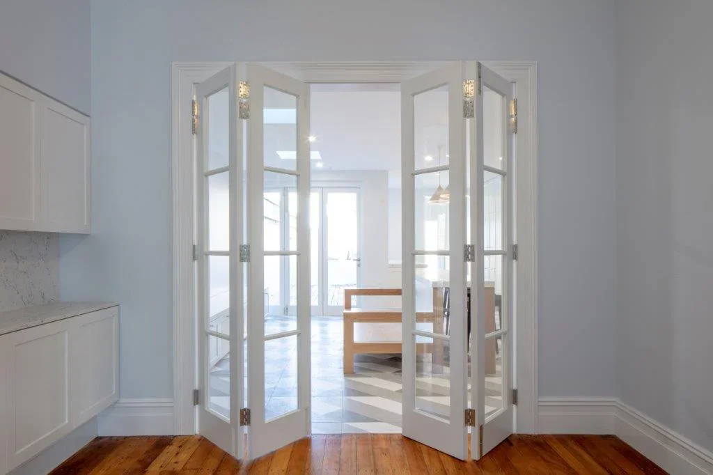 Open white double French doors leading to a bright dining area with a wooden table and black and white patterned floor.