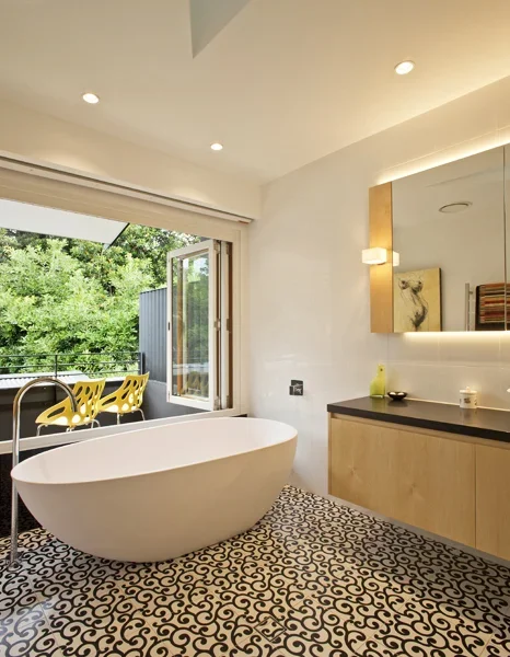 Modern bathroom with a freestanding bathtub, open window to a balcony with yellow chairs, and a vanity with a mirror and artwork.