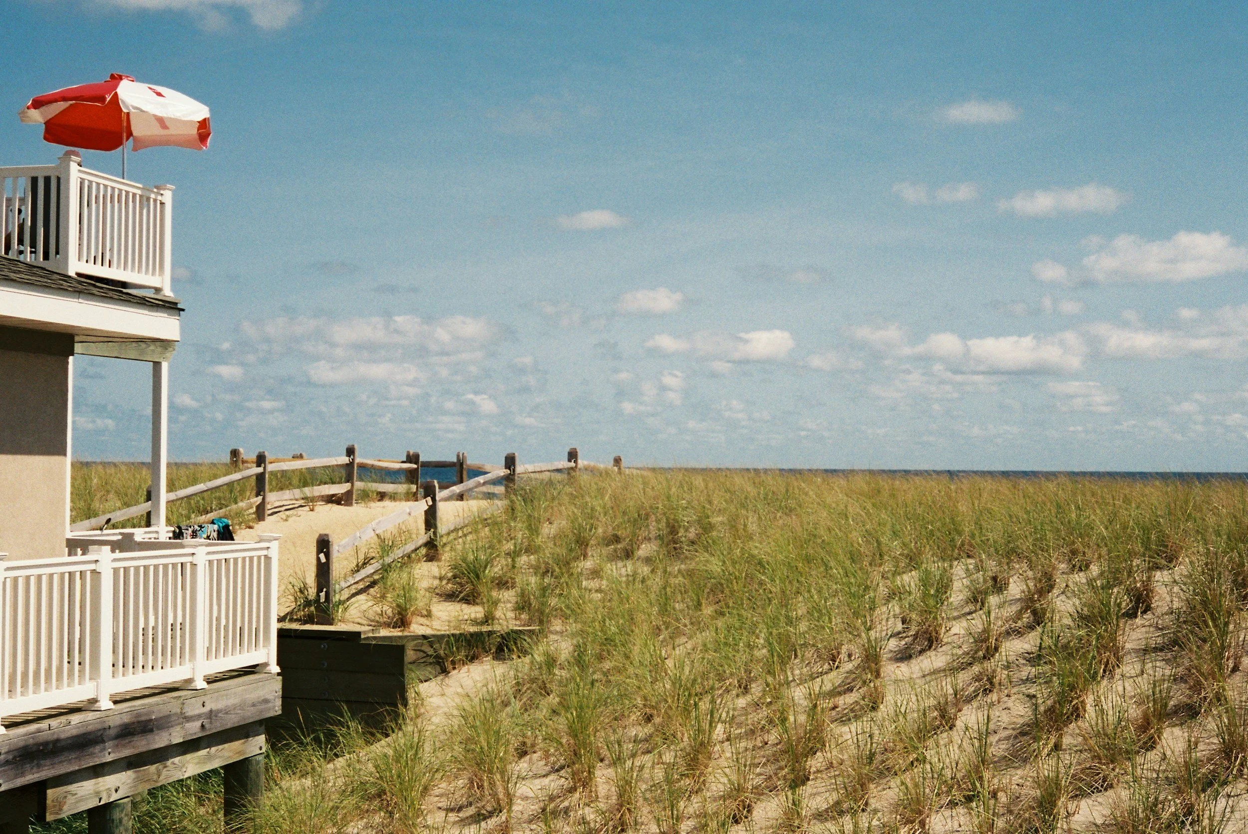 Beach house with white railings and a red and white umbrella on a balcony overlooking grassy dunes and the ocean under a blue sky with scattered clouds.