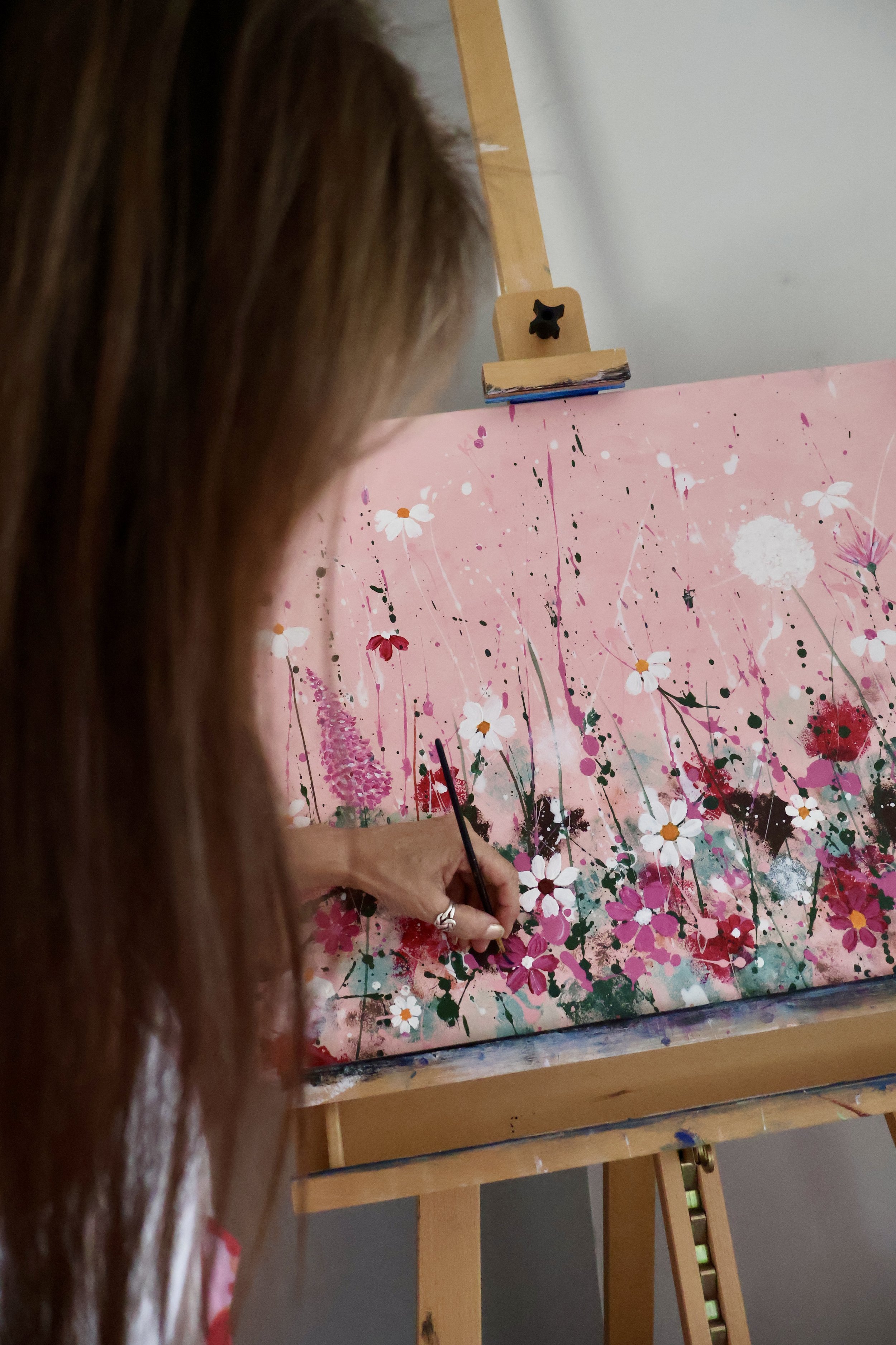 A woman painting a floral abstract artwork on a pink canvas, with her hand holding a thin paintbrush.