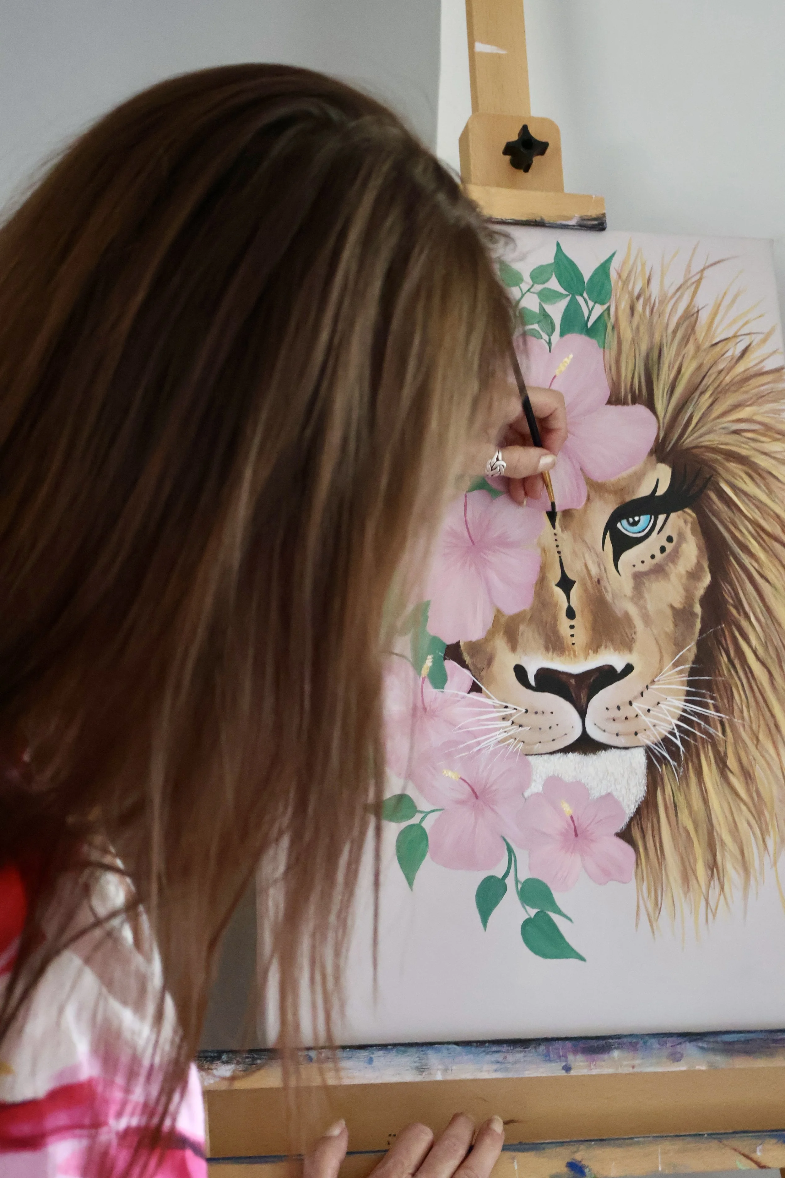 An artist painting a colorful lion with a pink floral mane on a canvas.