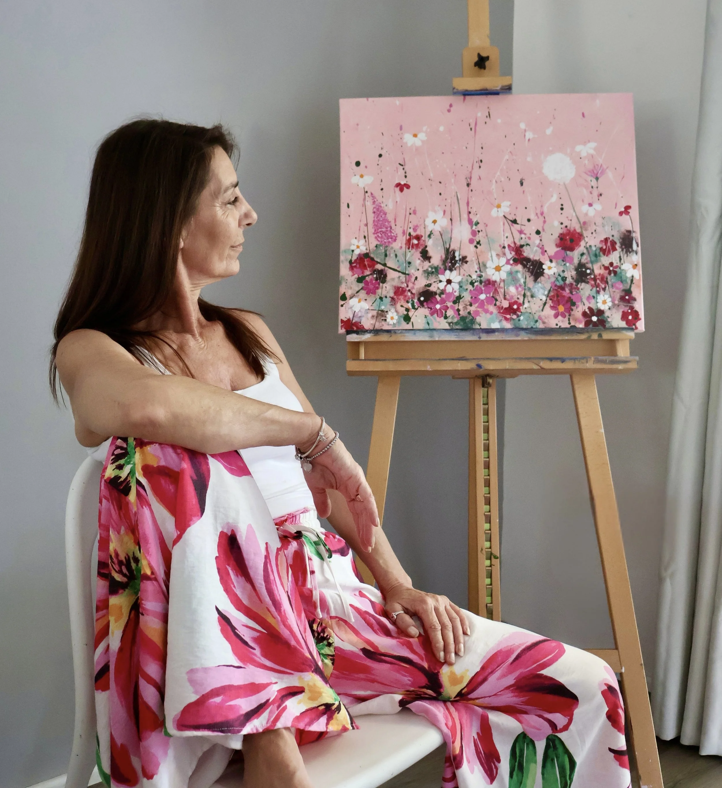 A woman sitting on a white chair gazing at her floral painting on an easel, wearing a matching floral skirt and top.