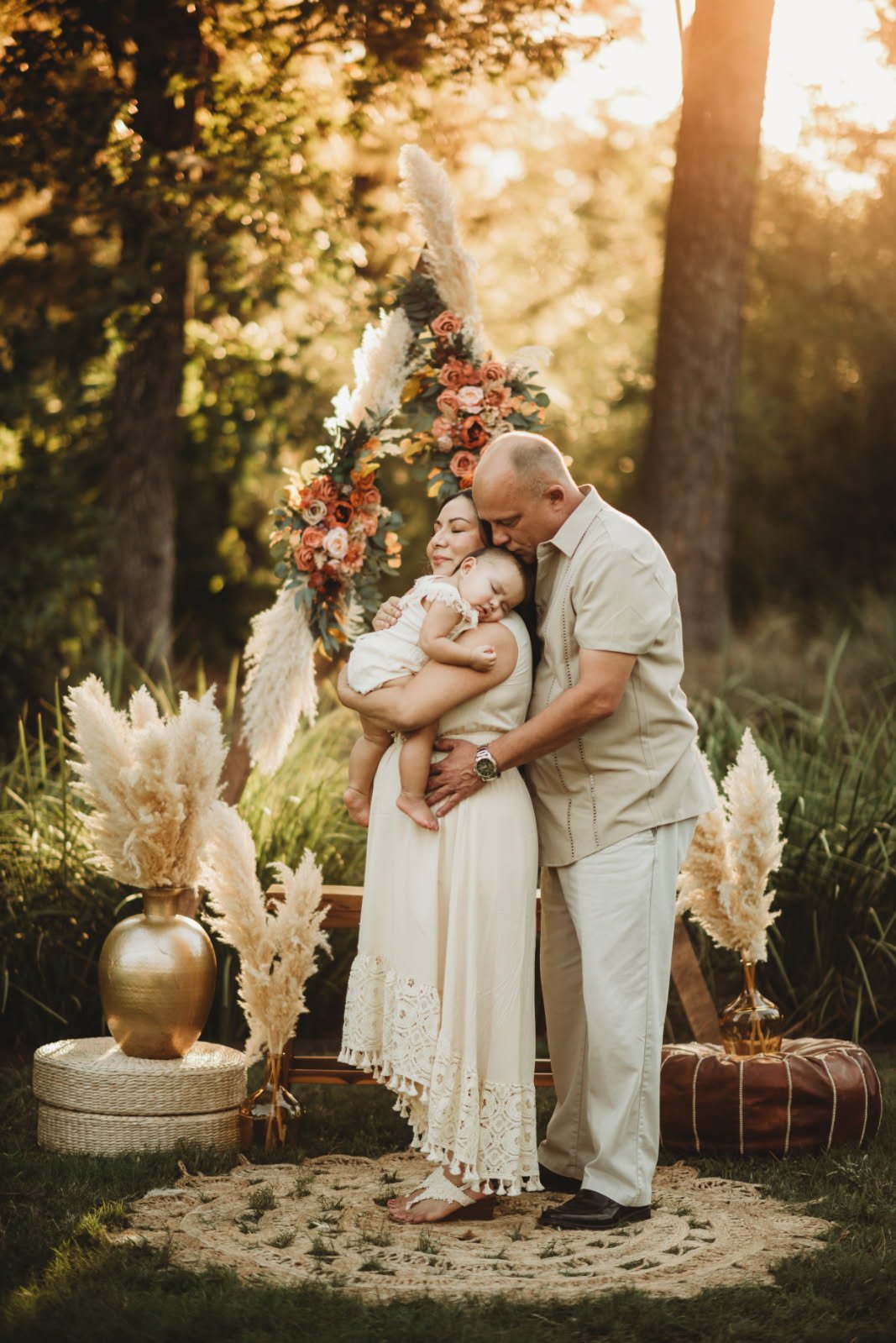 A family of three standing outdoors during sunset, embracing each other. The woman holds a sleeping child, and the man kisses her forehead. They are surrounded by decorative pampas grass and flowers, with trees and sunlight in the background.