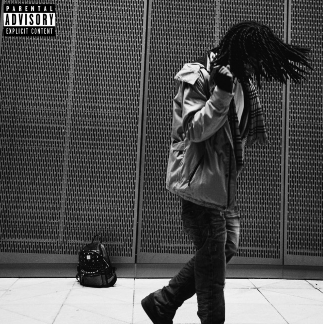 A person with braids, wearing a jacket and jeans, standing against a perforated wall, holding a phone to their ear with a backpack on the floor beside them.