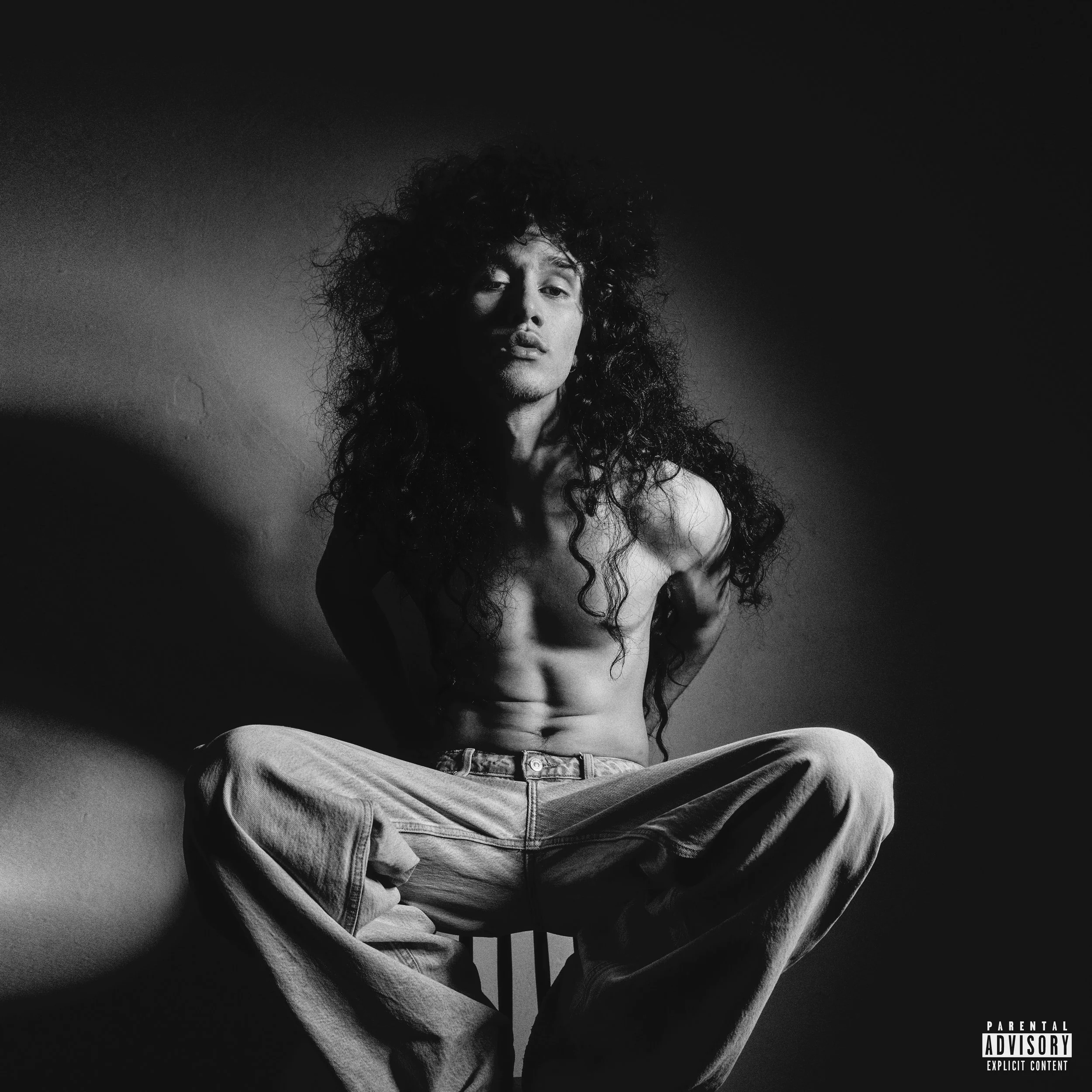 Black and white photo of a shirtless man with long, curly hair sitting cross-legged against a plain wall.