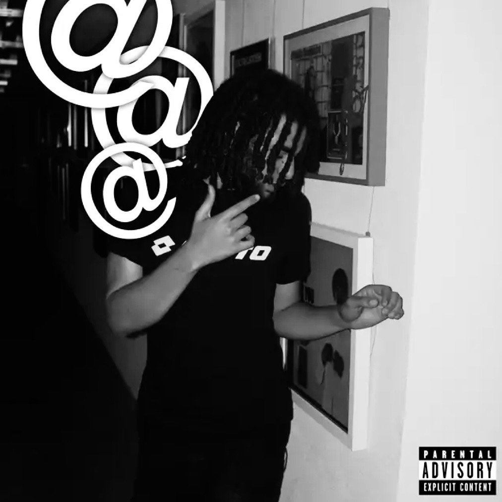 A young man with dreadlocks and glasses standing in front of a wall with framed pictures, making a gesture with his right hand near his face, while holding an object in his left hand. A large 'at' symbol graphic is overlaid on the left side of the image.