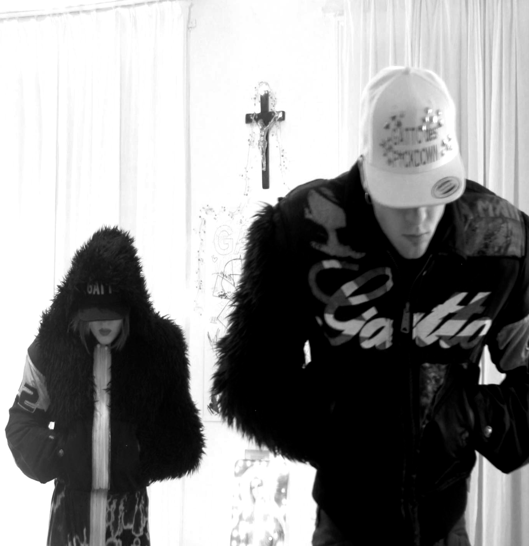 A man and a woman in fashionable winter clothing, with the woman in a fur-lined hood. They are in a room with white walls and a crucifix on the wall, and there are drawings on the wall behind them.