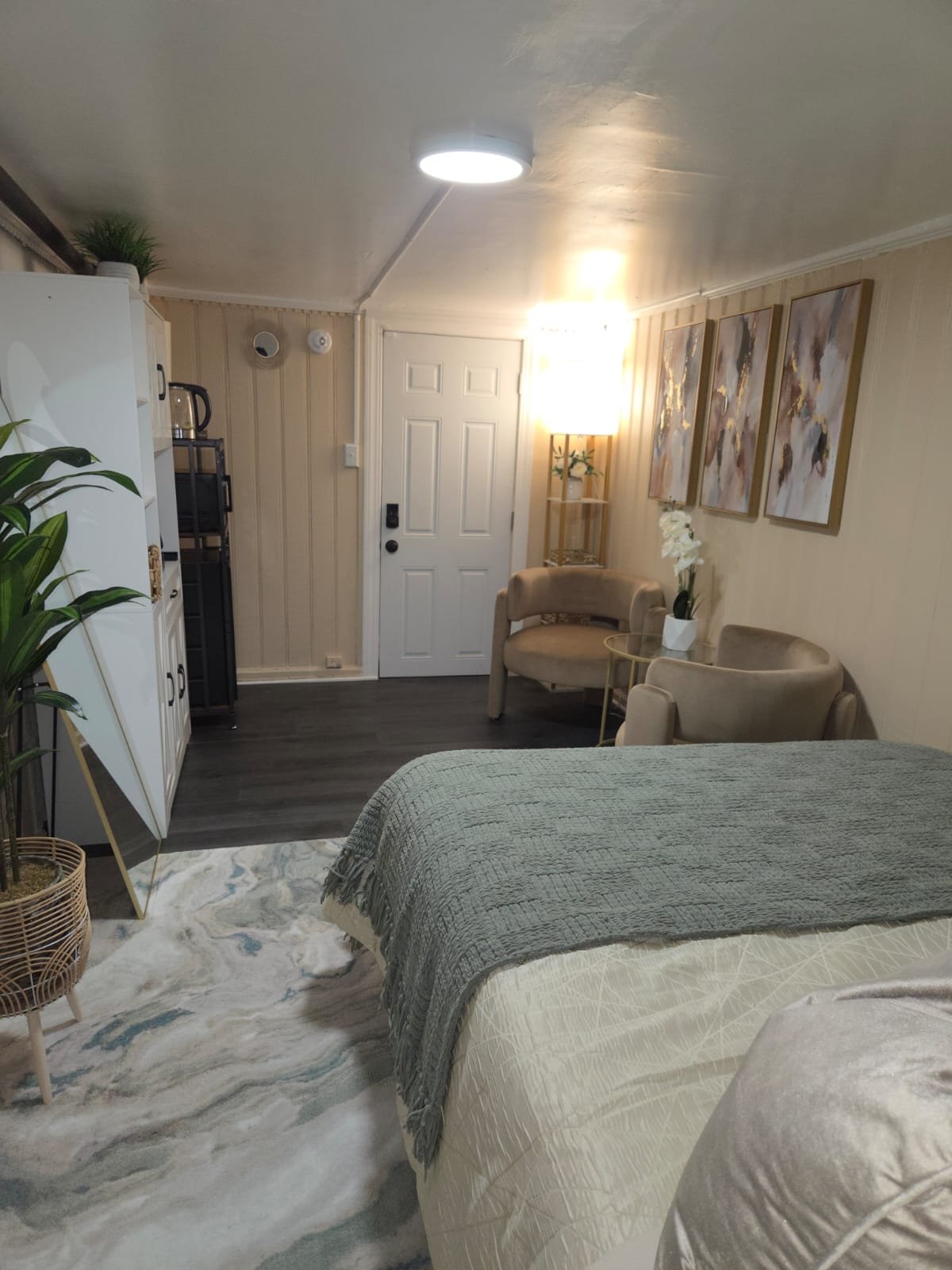 View of a cozy hotel room with a bed, a sitting area with two chairs and a small round table, wall art, a lamp, and a potted plant, with an entry door in the background.