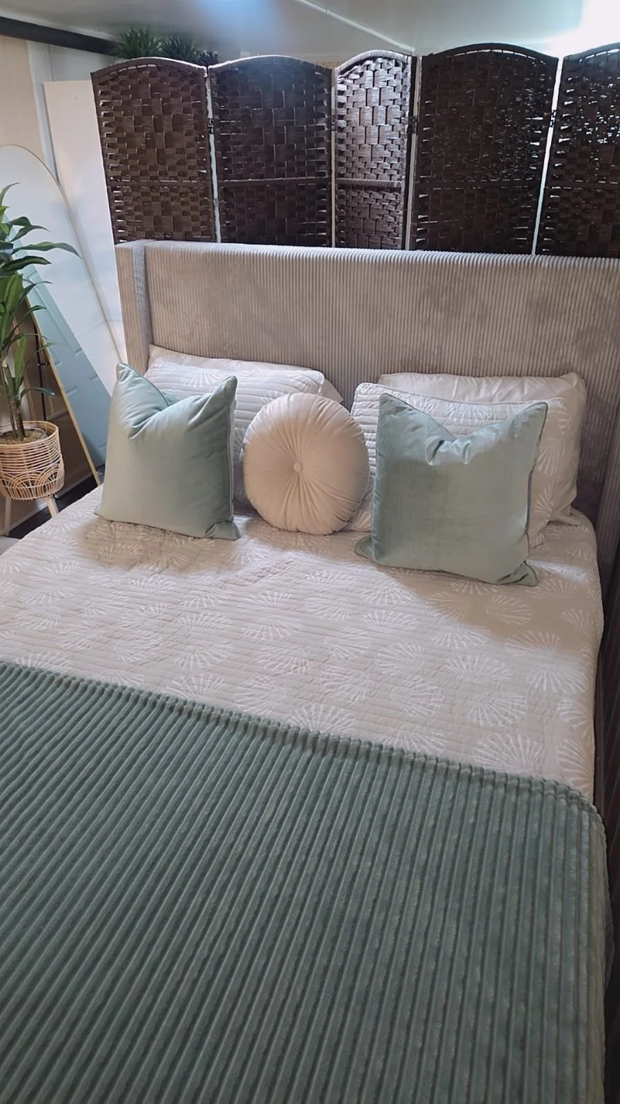 A cozy bedroom with a beige upholstered headboard, pillows, and a green blanket. Behind the bed is a decorative brown wooden privacy screen. There is a mirror and potted plant on the side.
