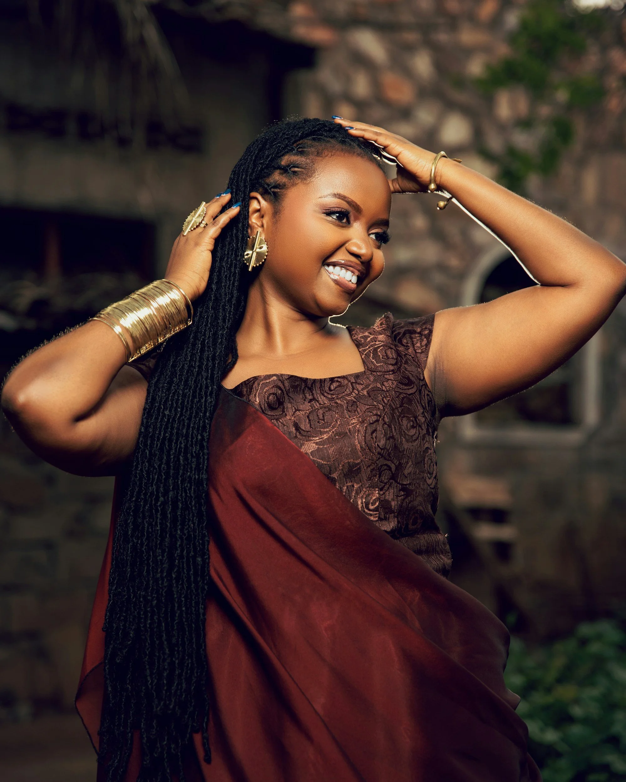A woman with long black braided hair smiling and posing with her hands on her head, wearing gold jewelry, a brown and maroon dress, outdoors with a stone building in the background.
