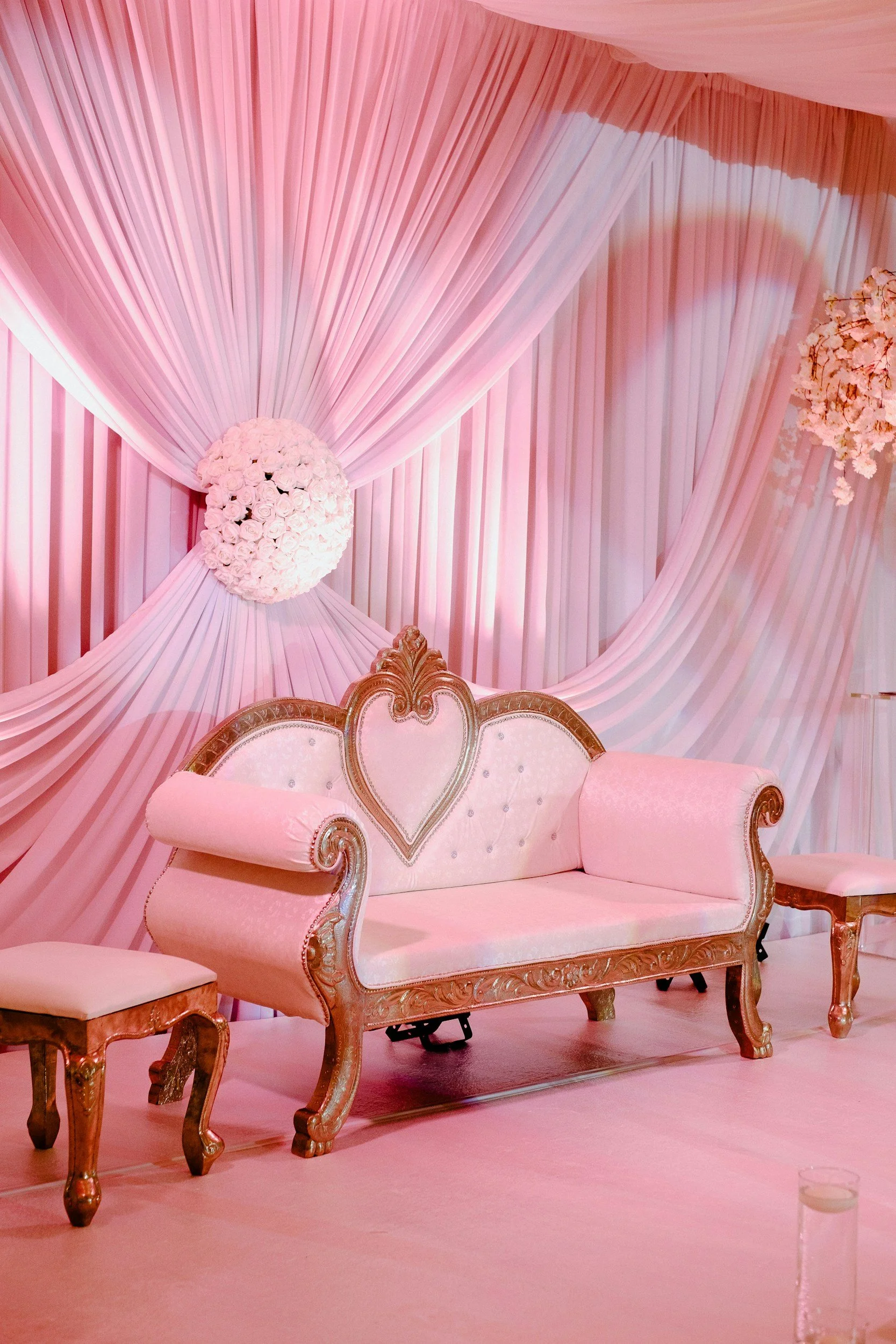 An elegant pink pastels wedding or event backdrop with draped fabric, a floral centerpiece, and ornate vintage-style furniture including a sofa and stools.