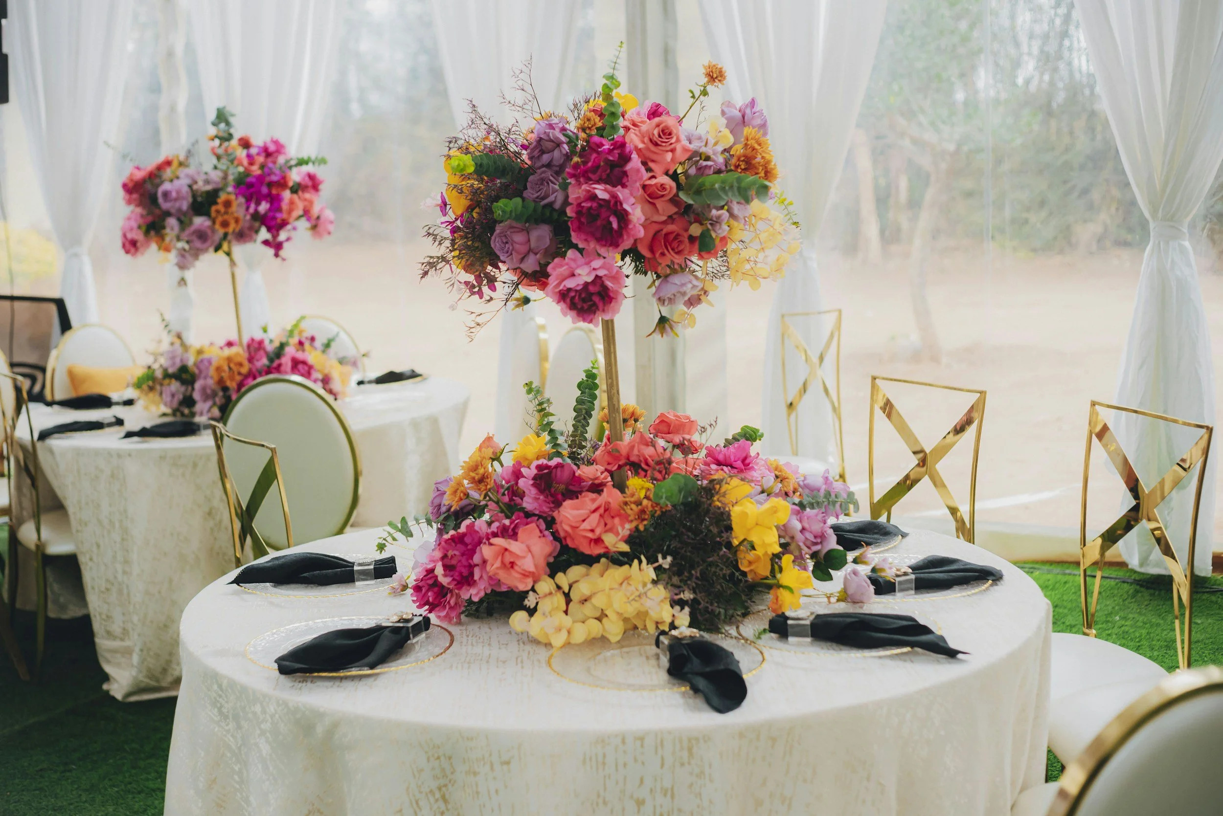 Round table decorated with a large floral centerpiece at a wedding or event reception, with white tablecloth and black napkins in a decorated outdoor venue with white drapes.