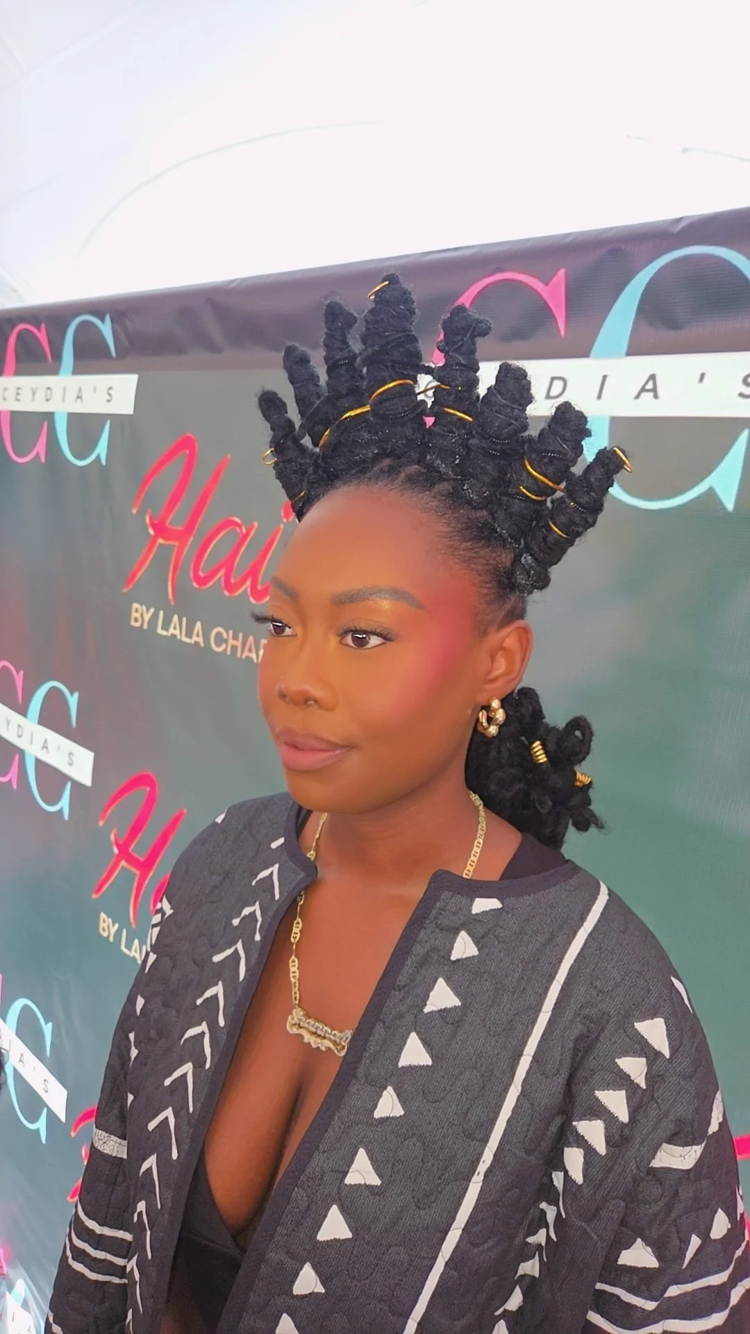 A woman with twisted bantu knots styled with gold hair rings, wearing gold jewelry, and a patterned gray jacket, standing in front of a colorful backdrop with text.
