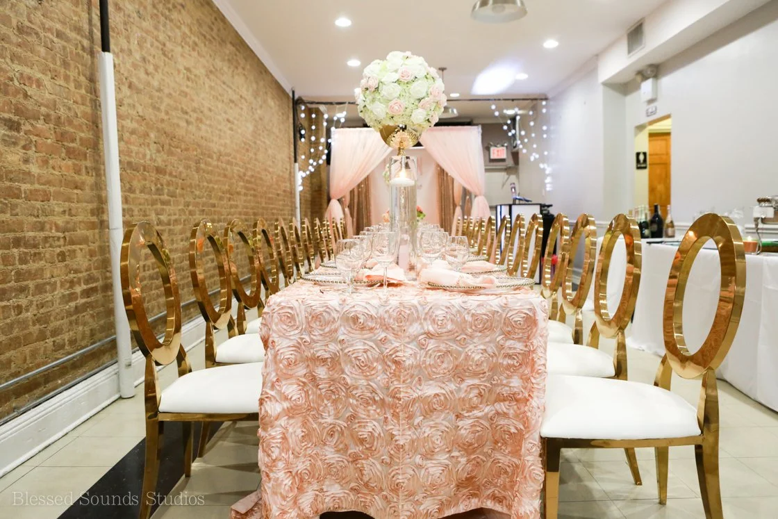 Elegant dining table set for a wedding or special celebration, featuring a pink floral tablecloth, a tall floral centerpiece with white and pink flowers, and gold chairs with white cushions in a decorated indoor venue with string lights and pink curtains.