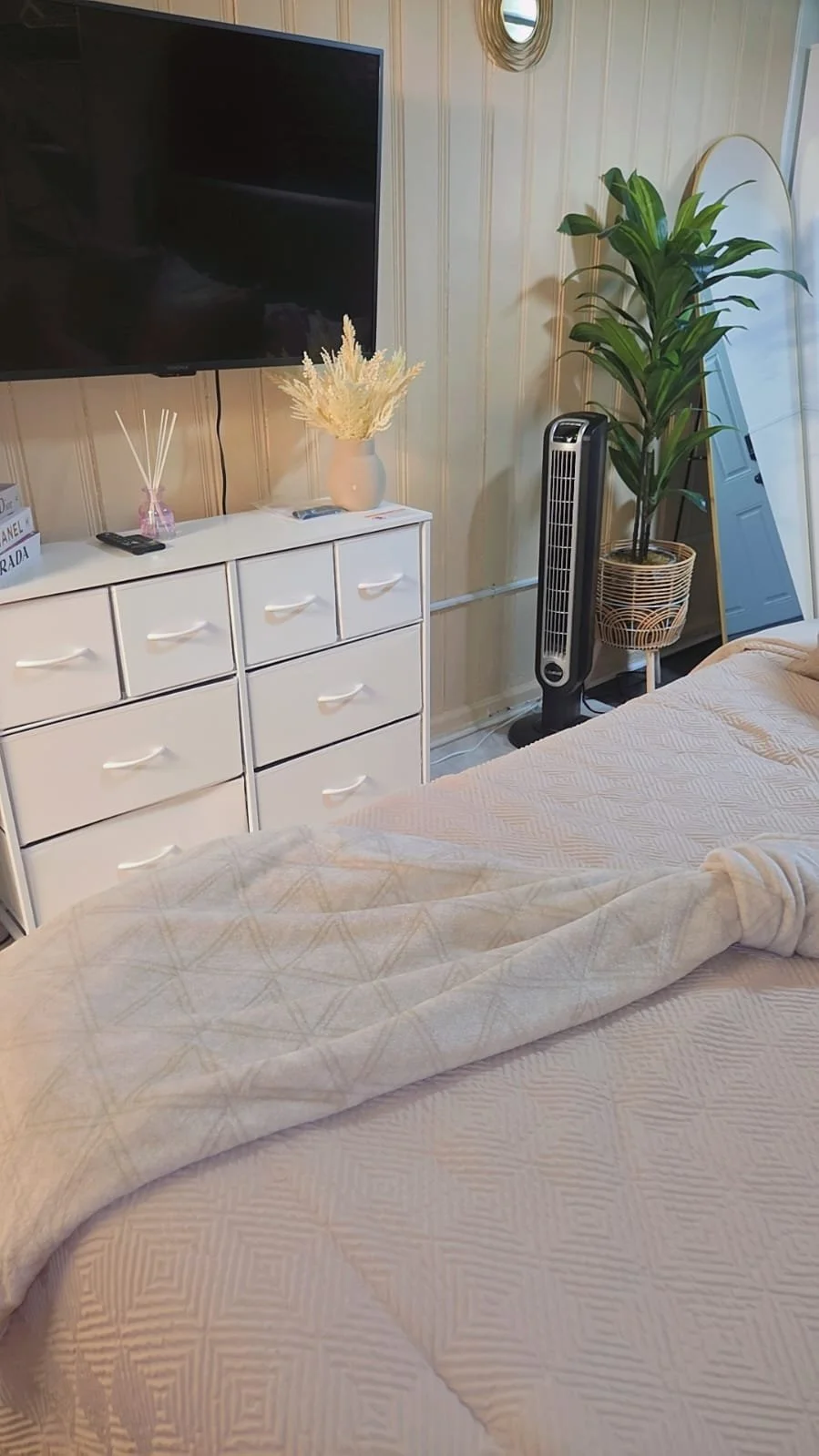 Bedroom with a white dresser, wall-mounted TV, decorative white vase with dried plants, small reed diffuser, remote control, large leafy potted plant, desk fan, upright mirror, and bed with light-colored quilted blanket.