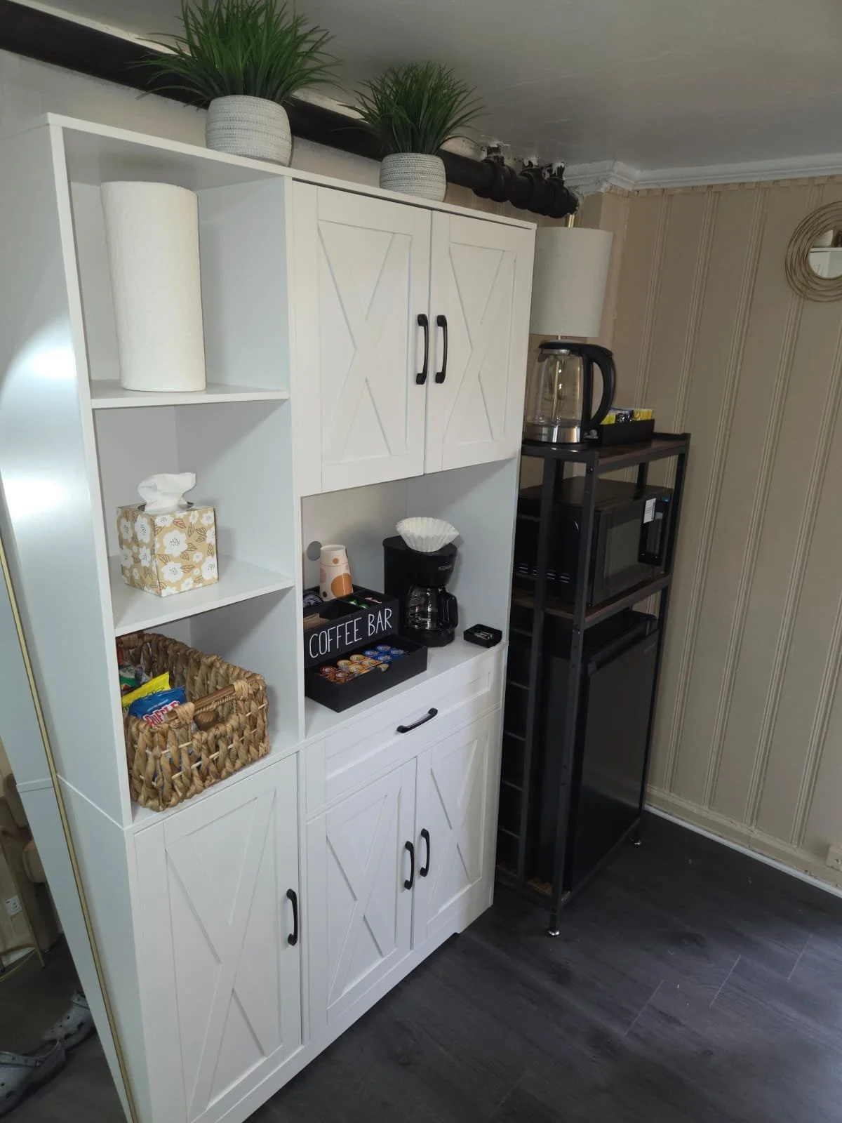 Coffee station with white cabinet, black hardware, coffee maker, glass tea kettle, black shelves, paper towel roll, tissue box, decorative plants on top, and striped wallpaper in the background.
