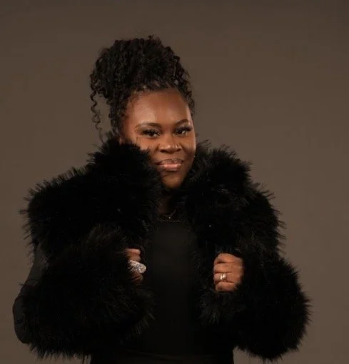 A woman with curly hair in a high ponytail, wearing a black outfit and a black fur coat, posing confidently against a plain background.