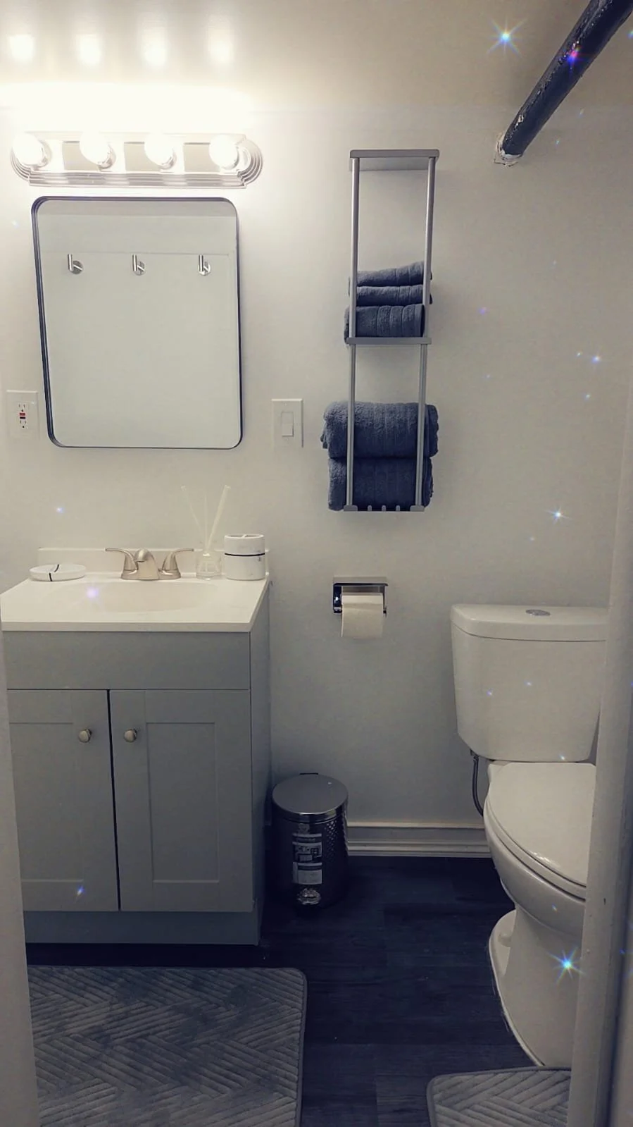 A small modern bathroom with white walls, a white vanity with a sink, a mirror above, a set of three towel hooks, a wall-mounted towel rack with blue towels, a toilet, and a small trash can. The room has dark wood flooring and two small gray rugs.