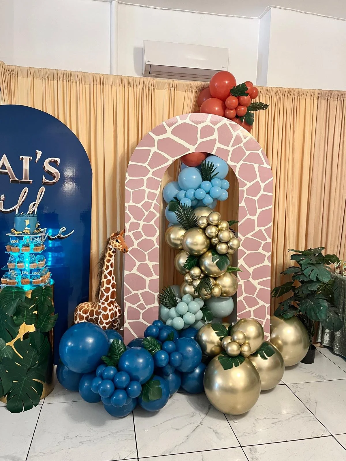 Decorative balloon arch with pink giraffe-patterned archway, gold, blue, and red balloons, tropical leaves, a giraffe plush toy, and a blue cupcake tower.