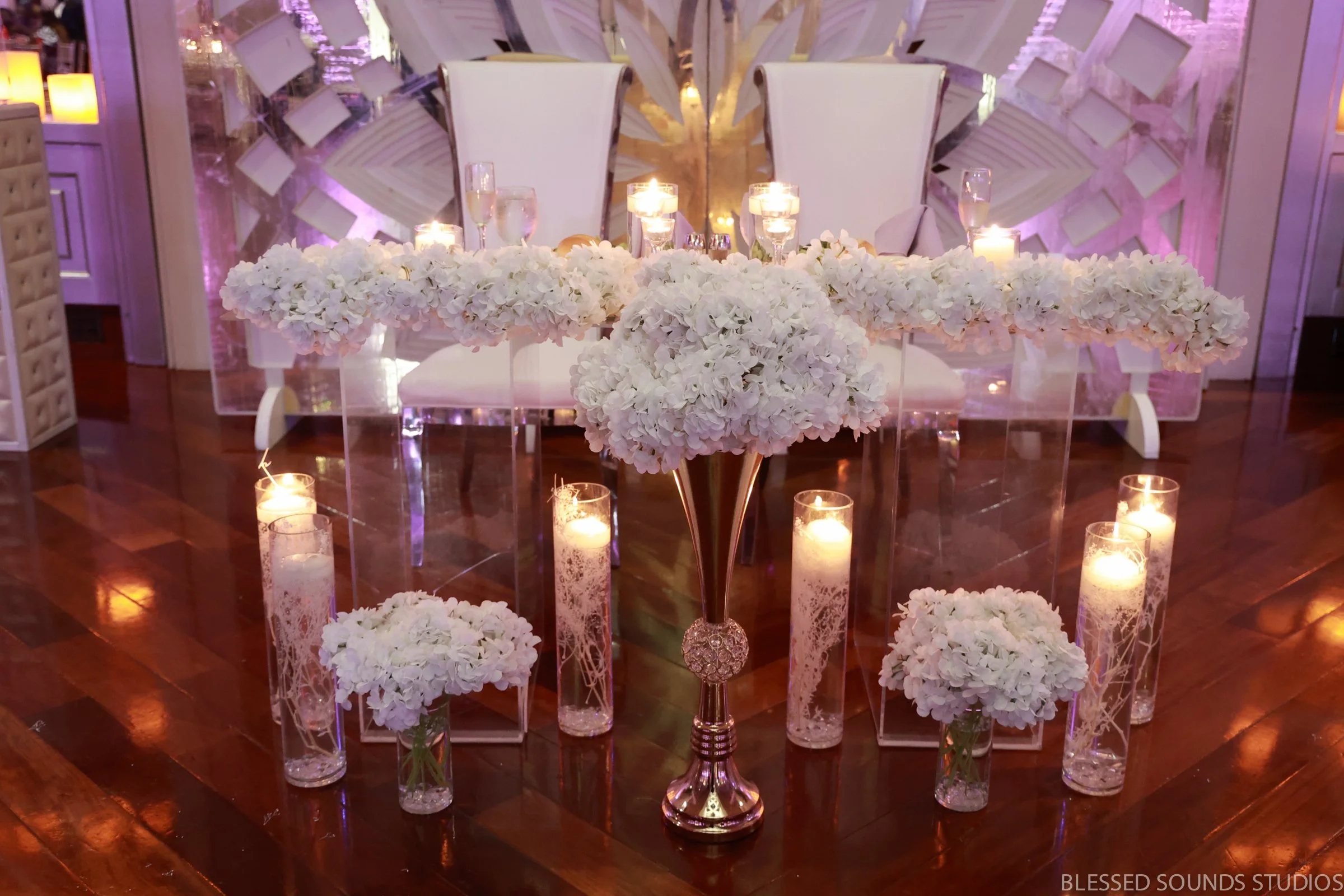 Elegant wedding table decorated with white floral arrangements, candles, and glassware. White chairs are positioned behind the table on a wooden floor, with a decorative white and purple background.