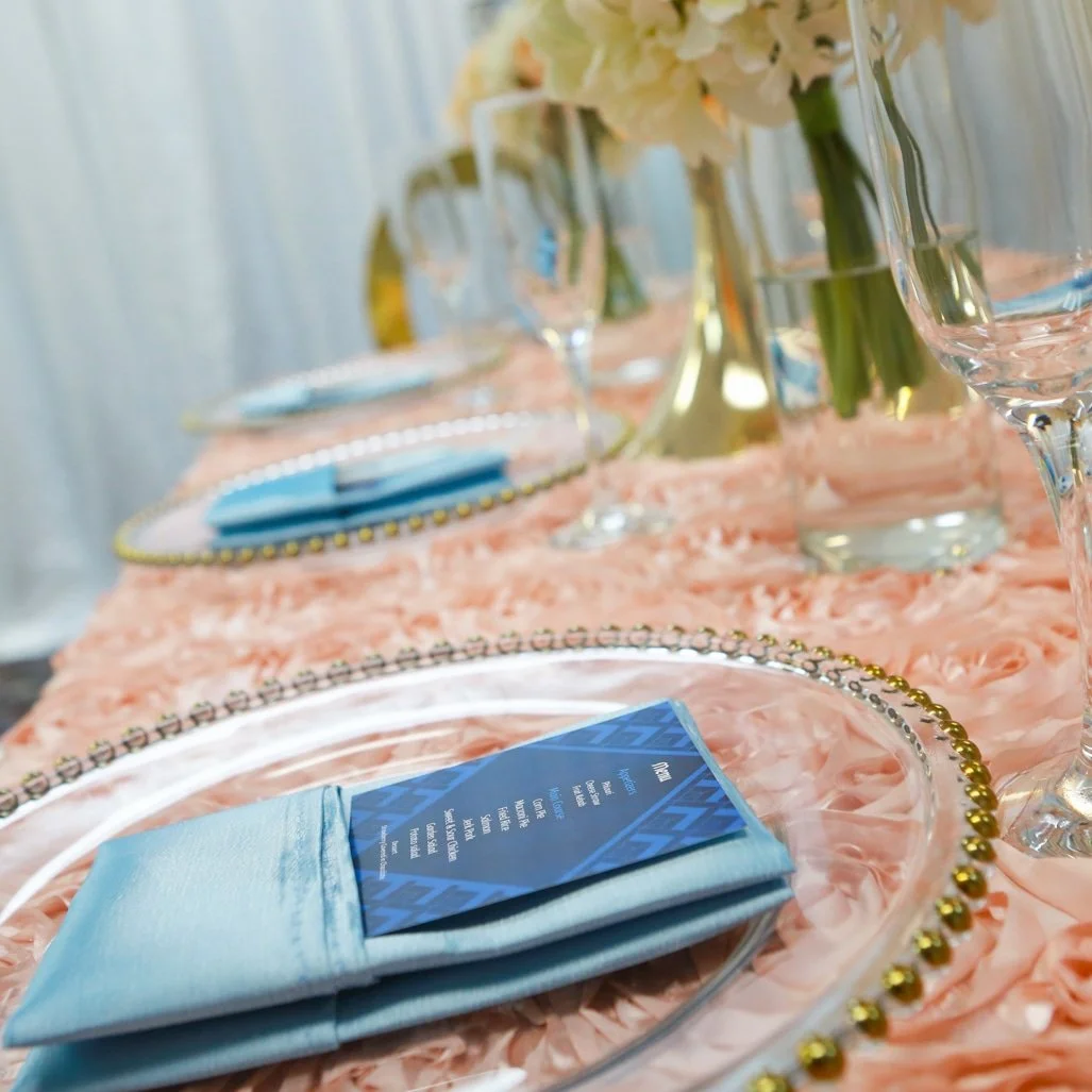 Elegant table setting with pink tablecloth, glass plates, blue napkins, and menu cards. Centerpieces include white flowers in glass vases, with wine glasses arranged on the table.