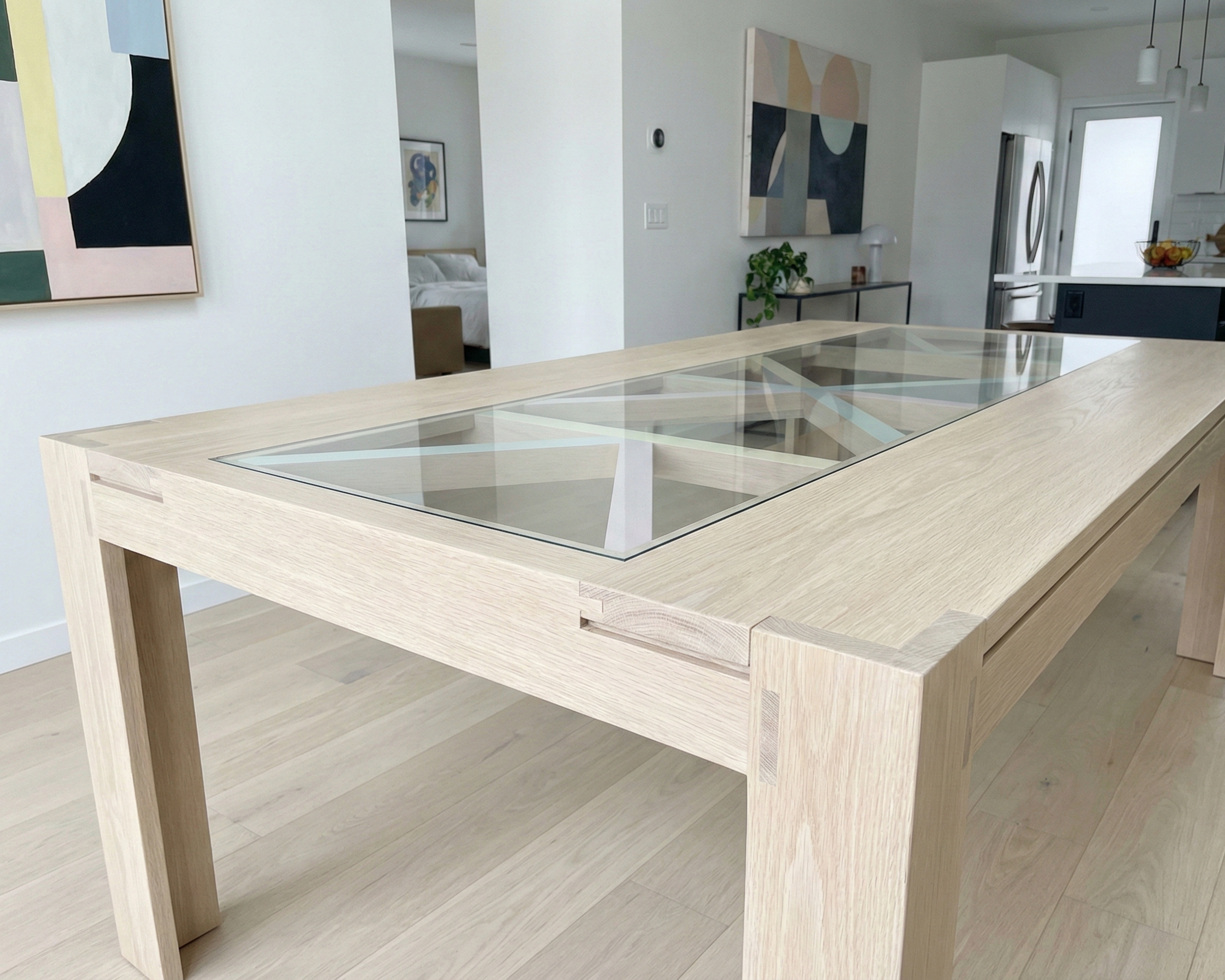 A modern dining table made of light wood with a glass top, situated in a bright, contemporary open-concept living space.