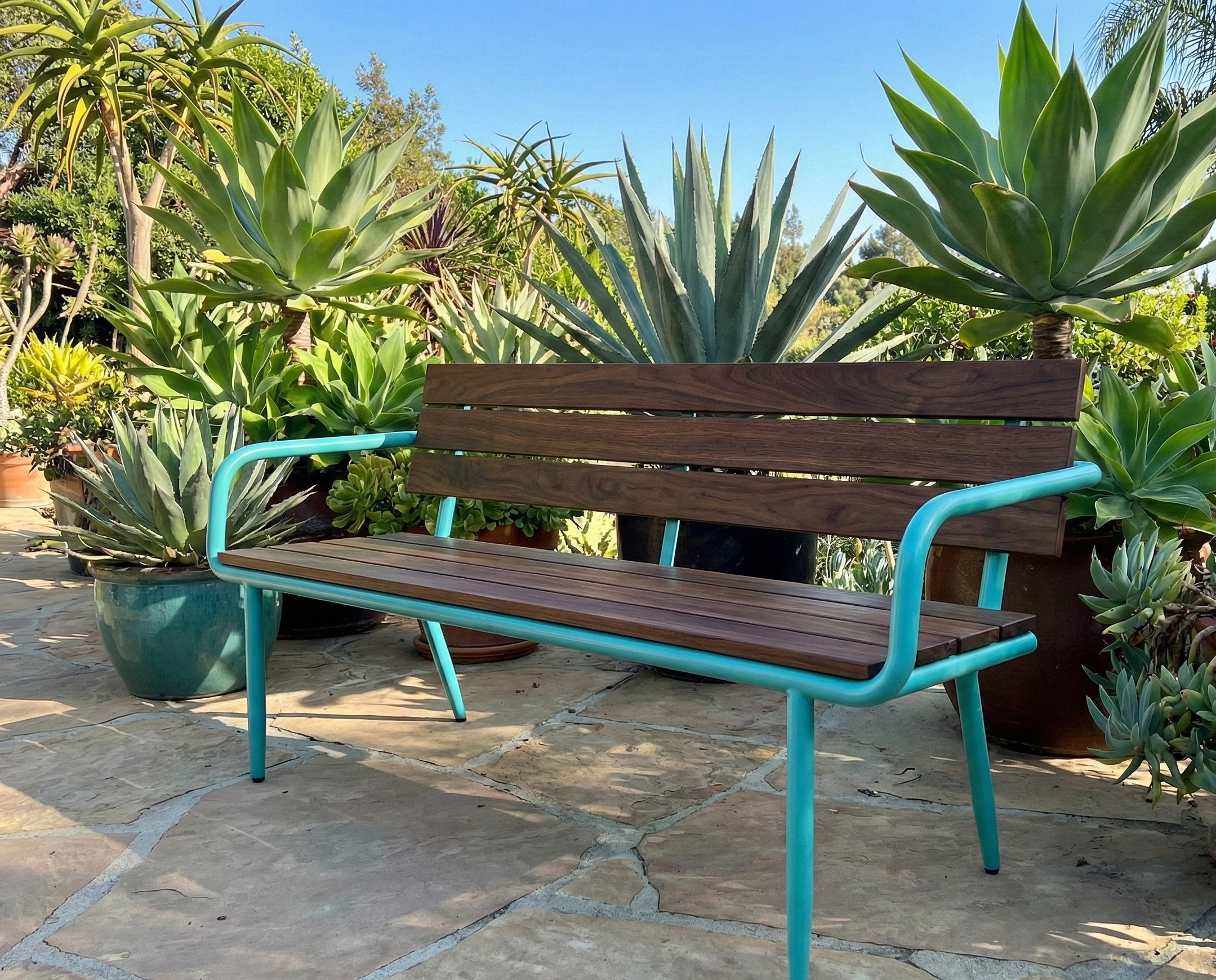 A wooden and turquoise metal bench on a stone patio surrounded by various potted succulents and large agave plants outdoors under a clear blue sky.