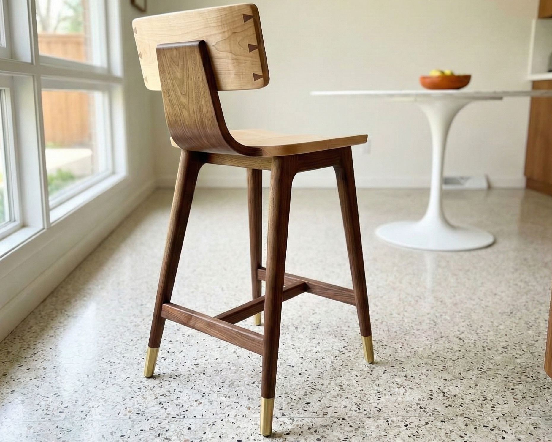A wooden chair with a backrest and gold-tipped legs in a room with a large window and a white table with a bowl of fruit.