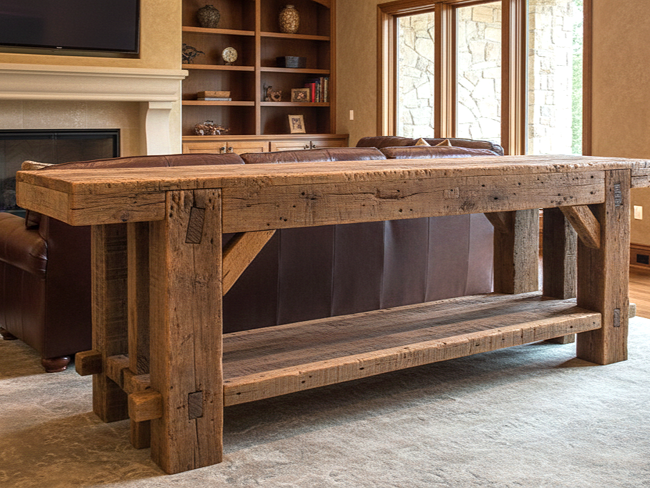 Rustic wooden dining or console table in a living room with large windows, brown leather furniture, a fireplace, and built-in shelves.
