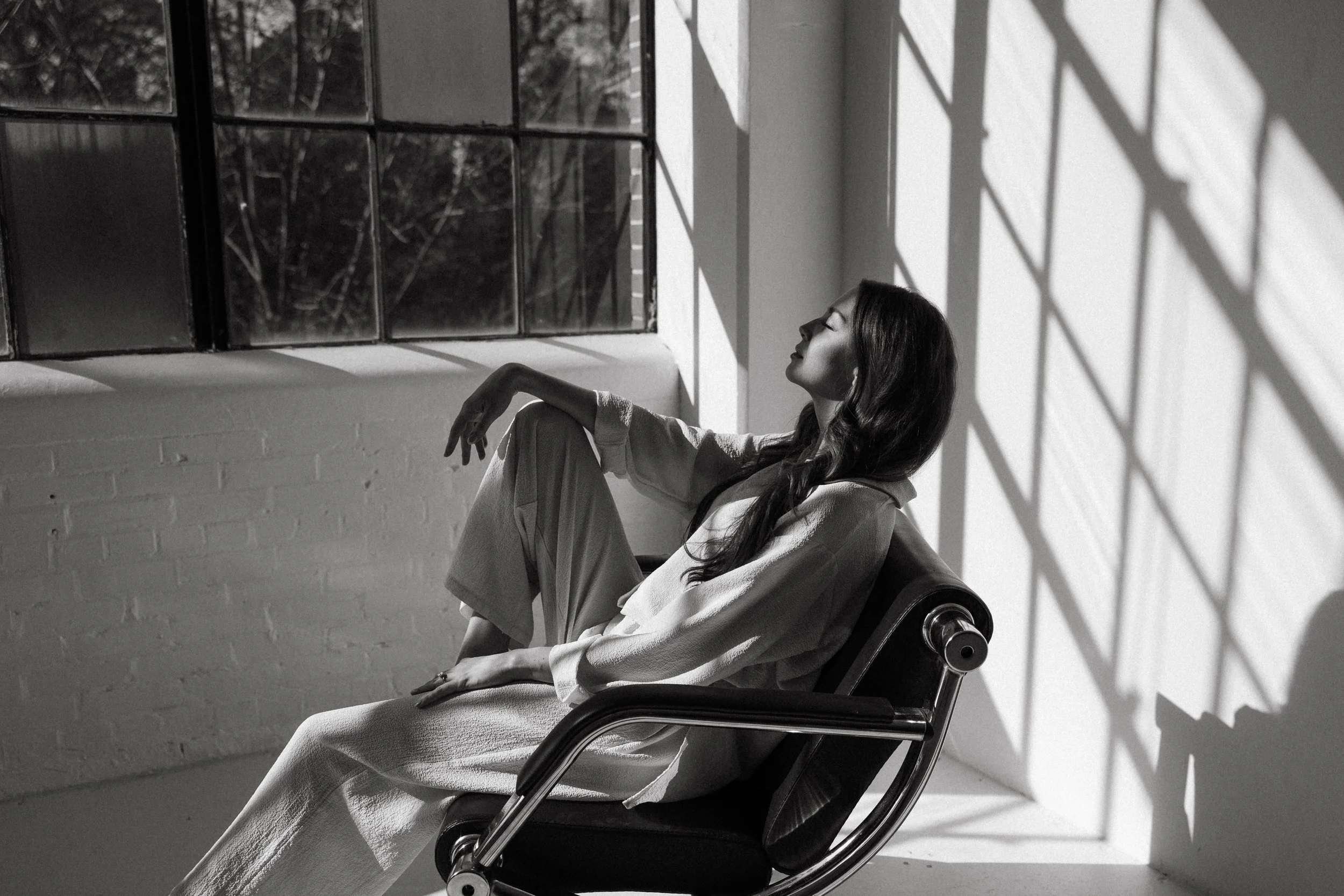 Black and white photo of a woman sitting in a chair near a window, with sunlight creating shadows on the wall.