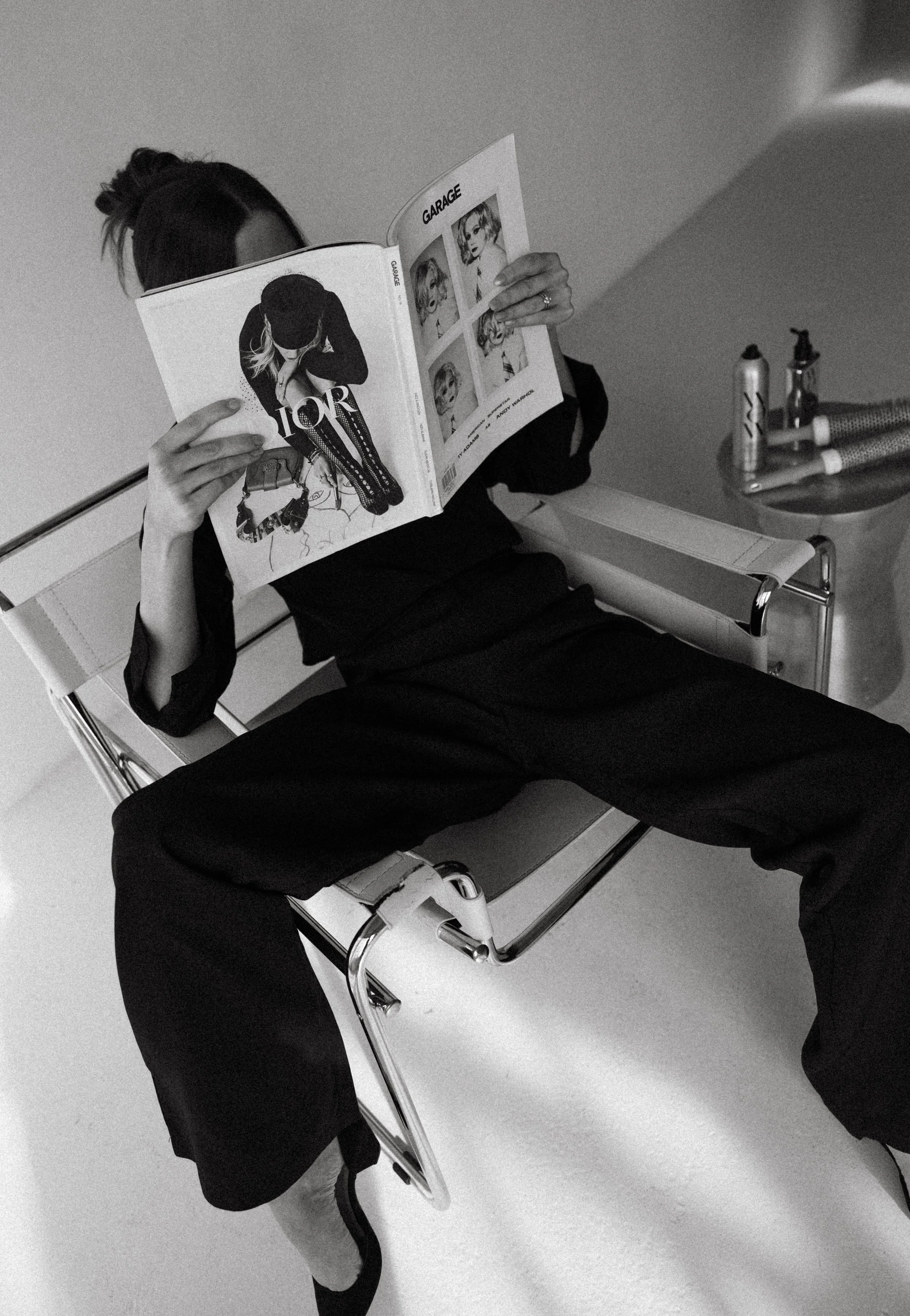 A woman wearing dark clothing and high heels is sitting on a chair, reading a magazine. A side table nearby holds a few cans and spray bottles.